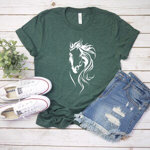 May include: A green t-shirt with a white silhouette of a horse's head. The shirt is laying on a wooden surface with a pair of white sneakers and a pair of blue denim shorts.