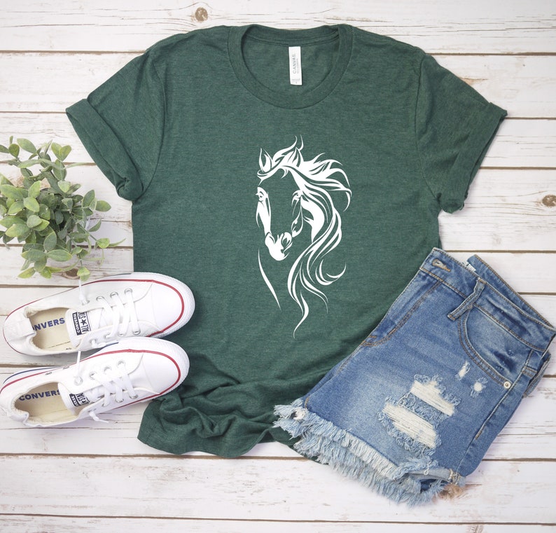 May include: A green t-shirt with a white silhouette of a horse's head. The shirt is laying on a wooden surface with a pair of white sneakers and a pair of blue denim shorts.