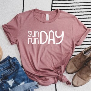 K&ouml;nnte beinhalten: Ein staubiges rosafarbenes T-Shirt mit dem Text "sun fun day" in wei&szlig;en Buchstaben. Das Shirt liegt flach auf einem wei&szlig;-schwarz gestreiften Teppich mit einem Paar braunen Wildlederstiefeln.