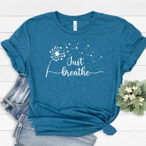 K&ouml;nnte beinhalten: Ein blaues T-Shirt mit einem wei&szlig;en L&ouml;wenzahnmuster und dem Text "Just breathe" auf der Vorderseite.