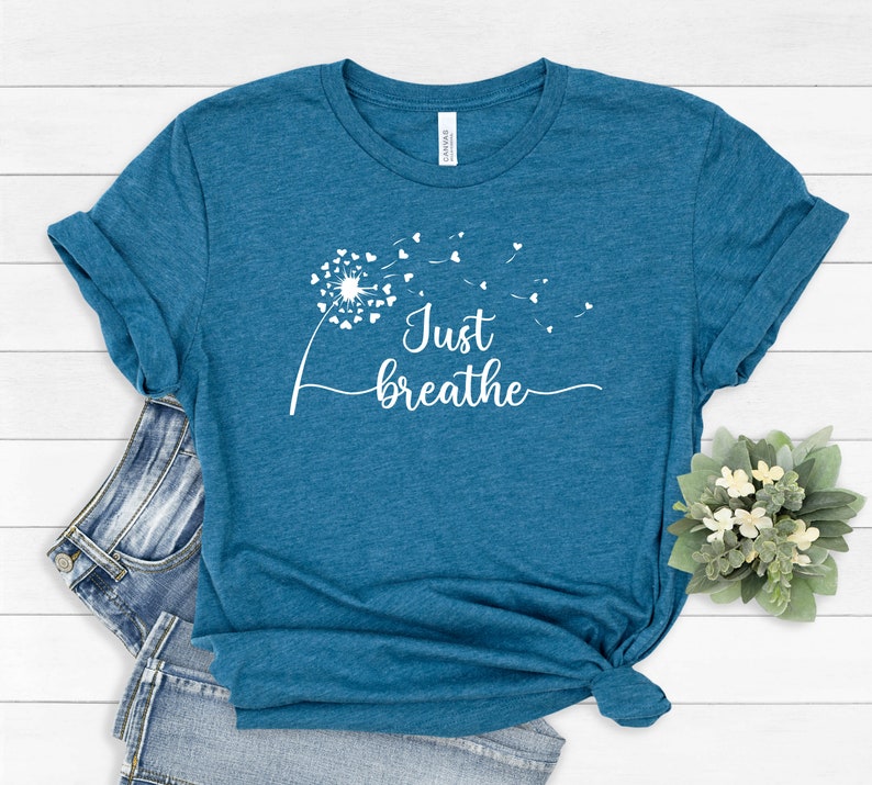K&ouml;nnte beinhalten: Ein blaues T-Shirt mit einem wei&szlig;en L&ouml;wenzahnmuster und dem Text "Just breathe" auf der Vorderseite.