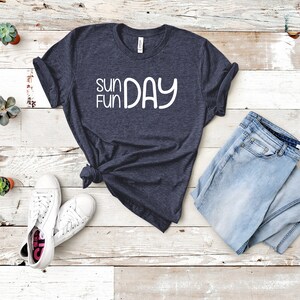 K&ouml;nnte beinhalten: Ein dunkelblaues T-Shirt mit dem Text "Sun Fun Day" in wei&szlig;en Buchstaben. Das Shirt liegt auf einer wei&szlig;en Holzoberfl&auml;che mit einem Paar wei&szlig;en Sneakers und einer blauen Jeans.