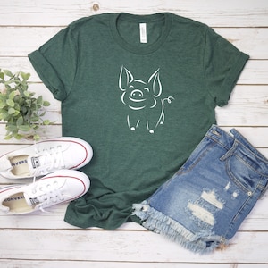 May include: A green t-shirt with a white outline of a pig on the front. The shirt is laying on a white wooden surface with a pair of white sneakers and a pair of blue denim shorts.