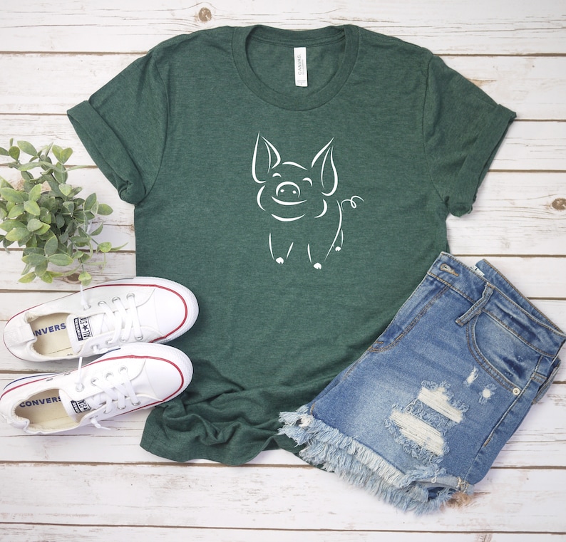 May include: A green t-shirt with a white outline of a pig on the front. The shirt is laying on a white wooden surface with a pair of white sneakers and a pair of blue denim shorts.