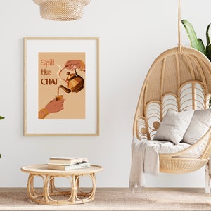 Op de afbeelding: Een ingelijst print met de tekst "Spill the Chai" in bruine en oranje letters. De print toont een hand die chai giet uit een bruine theepot in een glas. De print hangt aan een witte muur boven een bruine hangstoel van gevlochten riet.