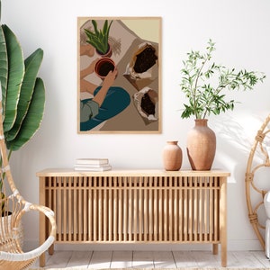 May include: A digital illustration of a person potting plants. The illustration is in a brown frame and hangs on a white wall. The person is wearing blue jeans and a light blue shirt. The person is holding a brown pot with a green plant in it. There are two brown paper bags with soil in them. The illustration is in a minimalist style.