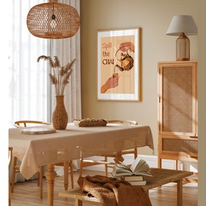 Op de afbeelding: Een lichtbruine rieten eettafel met een beige tafelkleed en een poster met de tekst "Spill the Chai" aan de muur. De poster toont een hand die chai uit een theepot in een kopje giet.
