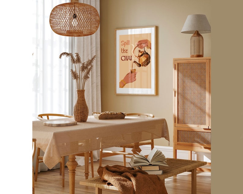 Op de afbeelding: Een lichtbruine rieten eettafel met een beige tafelkleed en een poster met de tekst "Spill the Chai" aan de muur. De poster toont een hand die chai uit een theepot in een kopje giet.