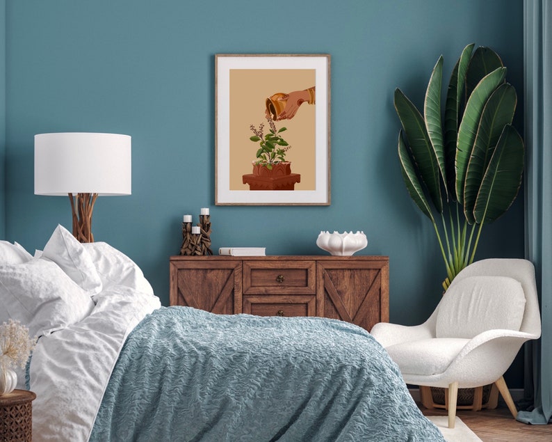 May include: A framed print of a hand watering a potted plant with a golden watering can. The print is on a light brown background and is framed in a simple black frame. The print is hanging on a blue wall.