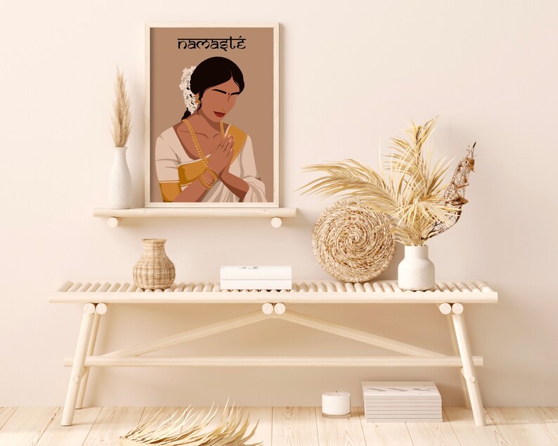 May include: A framed illustration of a woman in a white dress with gold jewelry, with her hands clasped in a prayer position. The background is brown and the text "namaste" is at the top of the frame. The illustration is on a white shelf with a white vase and a wicker vase on either side. A white bench with a wicker basket and a white vase with dried flowers is in front of the shelf.