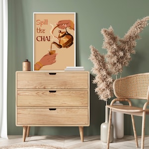 Op de afbeelding: Een houten dressoir met drie lades, een bruine keramische vaas met gedroogde bloemen en een ingelijst afdruk met de tekst "Spill the Chai" en een illustratie van een hand die chai giet uit een theepot in een glas.
