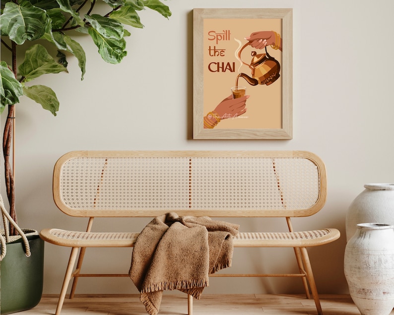 Op de afbeelding: Een ingelijst afdruk met een lichtbruine achtergrond en de tekst "Spill the Chai" in een bruin en oranje lettertype. De afbeelding toont een hand die chai uit een theepot in een glas giet.