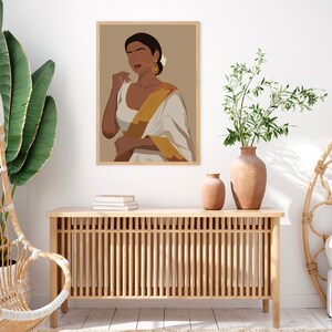 May include: A framed illustration of a woman wearing a white and gold sari with a brown background. The woman is looking to the side and has dark hair and brown eyes. She is wearing gold earrings and a nose ring.