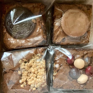 May include: Four brownies in clear plastic wrap, each with a different topping. One brownie has an Oreo cookie, another has a peanut butter cup, a third has chopped nuts, and the last has M&M candies.