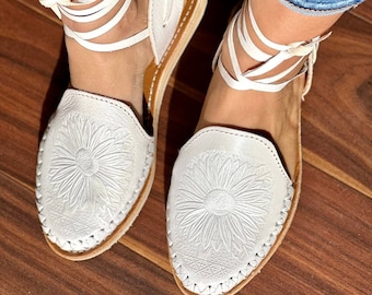 May include: White leather sandals with a sunflower design and white laces. The sandals have a brown sole and are worn on a wooden floor.