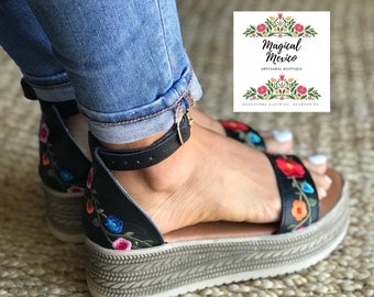 Könnte beinhalten: Schwarze Plateausandalen mit bunter Blumenstickerei. Die Sandalen haben eine dicke, gewebte Plateau-Sohle und einen Knöchelriemen. Die Sandalen befinden sich auf einem gewebten Teppich.