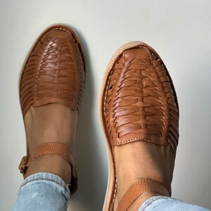 Pu&ograve; includere: Un paio di sandali in pelle marrone con cinturini intrecciati e cinturini alla caviglia. I sandali sono indossati con jeans blu.