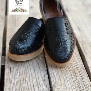 Peut inclure: Paire de huaraches en cuir noir avec des motifs floraux et de feuilles complexes. Les chaussures ont des d&eacute;tails tress&eacute;s sur les bords et des semelles beiges. Le logo "Magical Mexico" est visible.