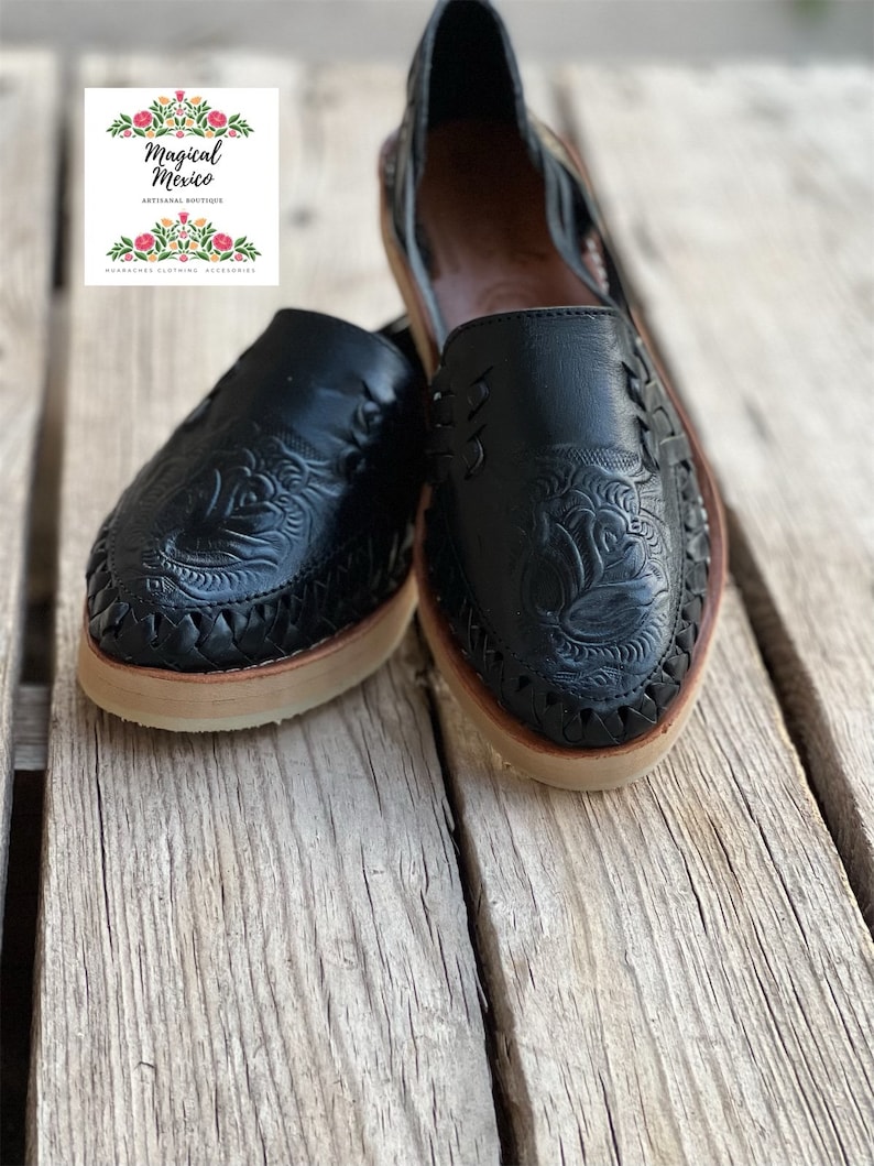 Peut inclure: Paire de huaraches en cuir noir avec des motifs floraux et de feuilles complexes. Les chaussures ont des d&eacute;tails tress&eacute;s sur les bords et des semelles beiges. Le logo "Magical Mexico" est visible.