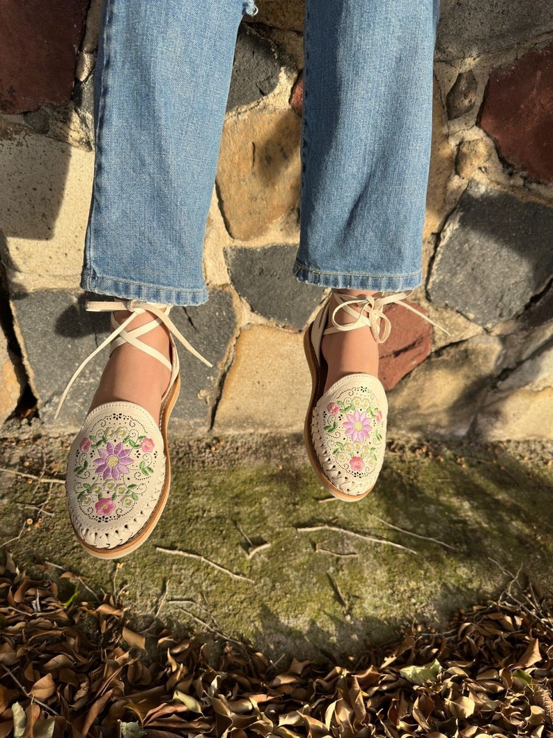 May include: Cream-colored leather sandals with floral embroidery in pink and purple. The sandals have a rounded toe and a woven design around the edges. They feature ankle ties and are worn with blue jeans.