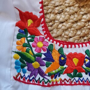 以下が含まれることがあります： 鮮やかな花柄刺繍が施された白いトップス。刺繍には、赤、黄、オレンジ、青、ピンク、紫の花と緑の葉が使われています。ネックラインは赤いステッチでトリミングされています。
