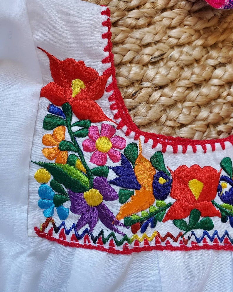 以下が含まれることがあります： 鮮やかな花柄刺繍が施された白いトップス。刺繍には、赤、黄、オレンジ、青、ピンク、紫の花と緑の葉が使われています。ネックラインは赤いステッチでトリミングされています。