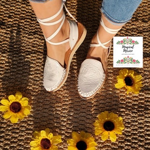K&ouml;nnte beinhalten: Wei&szlig;e Ledersandalen mit wei&szlig;en Schn&uuml;rsenkeln und Blumenstickerei. Die Sandalen befinden sich auf einer geflochtenen Strohmatte mit gelben Sonnenblumen.
