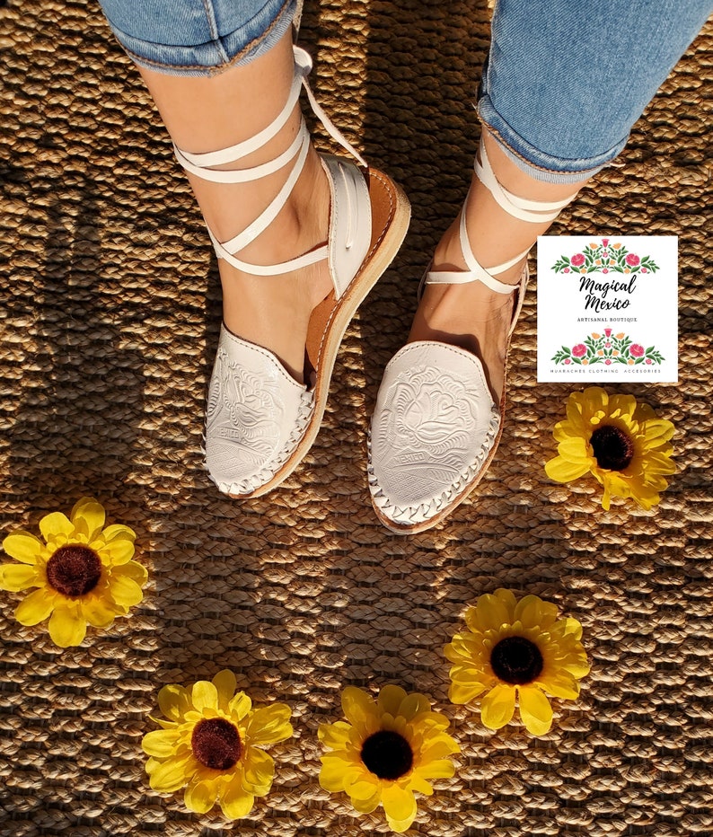 K&ouml;nnte beinhalten: Wei&szlig;e Ledersandalen mit wei&szlig;en Schn&uuml;rsenkeln und Blumenstickerei. Die Sandalen befinden sich auf einer geflochtenen Strohmatte mit gelben Sonnenblumen.