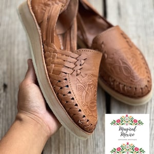 Peut inclure: Paire de huaraches en cuir marron avec des motifs floraux tiss&eacute;s et gaufr&eacute;s. Les chaussures ont une semelle de couleur claire et sont pr&eacute;sent&eacute;es sur une surface en bois. L'image comprend un logo avec le texte "Magical Mexico ARTISANAL BOUTIQUE HUARACHES CLOTHING ACCESORIES."