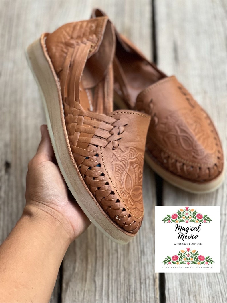 Peut inclure: Paire de huaraches en cuir marron avec des motifs floraux tiss&eacute;s et gaufr&eacute;s. Les chaussures ont une semelle de couleur claire et sont pr&eacute;sent&eacute;es sur une surface en bois. L'image comprend un logo avec le texte "Magical Mexico ARTISANAL BOUTIQUE HUARACHES CLOTHING ACCESORIES."