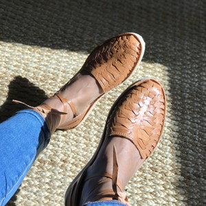 Puede incluir: Un par de sandalias de cuero marr&oacute;n con correas y cordones tejidos. Las sandalias se llevan en los pies de una persona, con los cordones atados alrededor de los tobillos.