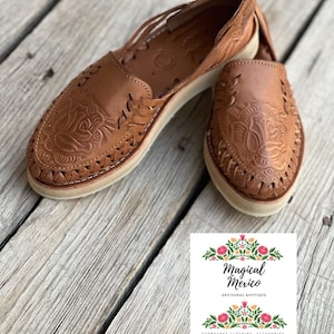 Peut inclure: Paire de huaraches en cuir marron avec gaufrage floral et d&eacute;tails tress&eacute;s. Les chaussures ont une semelle couleur cr&egrave;me. L'image comprend un logo avec le texte "Magical Mexico" et des motifs floraux.