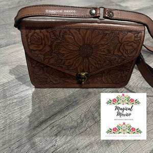 May include: Brown leather crossbody bag with floral and sunflower embossed design. The bag features a flap closure and adjustable strap. The image also includes a small card with the text "Magical Mexico ARTISANAL BOUTIQUE HUARACHES CLOTHING ACCESSORIES".