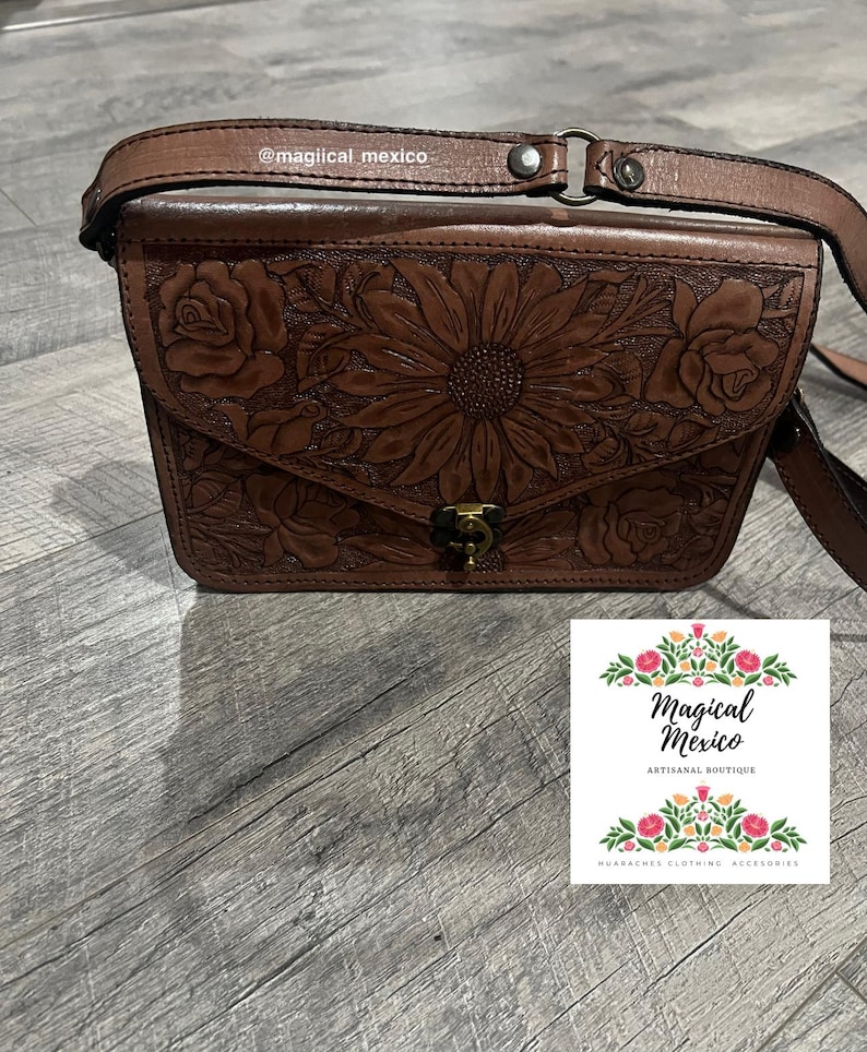 May include: Brown leather crossbody bag with floral and sunflower embossed design. The bag features a flap closure and adjustable strap. The image also includes a small card with the text "Magical Mexico ARTISANAL BOUTIQUE HUARACHES CLOTHING ACCESSORIES".