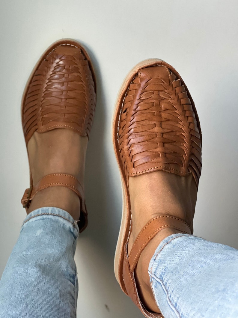Pu&ograve; includere: Un paio di sandali in pelle marrone con cinturini intrecciati e cinturini alla caviglia. I sandali sono indossati con jeans blu.