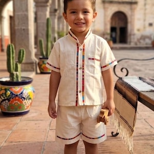 Puede incluir: Un conjunto de camisa de manga corta con botones y pantalones cortos a juego en color crema. La camisa presenta un cuello mao, un bolsillo en el pecho y un ribete vertical con un patrón tejido colorido. Los pantalones cortos tienen un ribete a juego.