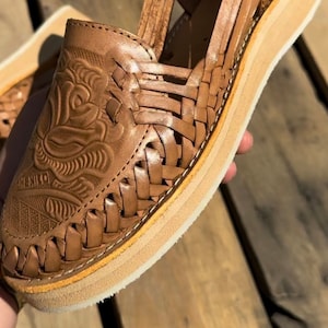 Peut inclure: Huaraches en cuir marron avec un motif tress&eacute; et une semelle plateforme. L'avant de la chaussure pr&eacute;sente des motifs floraux et g&eacute;om&eacute;triques en relief. La semelle mesure environ 2,5 cm d'&eacute;paisseur. Le mot "MEXICO" est estamp&eacute; sur la chaussure.