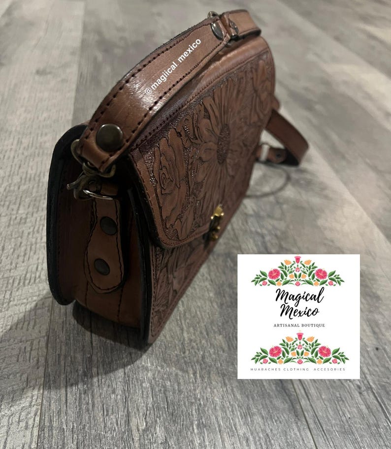 May include: Brown leather crossbody bag with embossed floral design. The bag features a flap closure with a gold-colored clasp and a long adjustable strap. The text "@magical mexico" is visible on the strap.