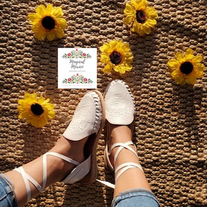 K&ouml;nnte beinhalten: Wei&szlig;e Ledersandalen mit aufwendigen Blumenstickereien und wei&szlig;en Schn&uuml;rsenkeln, die die Beine hochbinden. Die Sandalen befinden sich auf einem gewebten Teppich mit gelben Sonnenblumen.