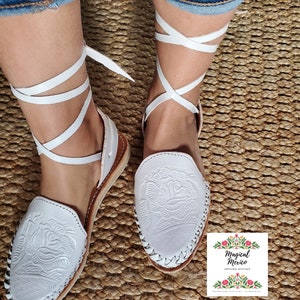 K&ouml;nnte beinhalten: Wei&szlig;e Ledersandalen mit Rosenmuster und wei&szlig;en Schn&uuml;rsenkeln, die sich um die Kn&ouml;chel binden. Die Sandalen befinden sich auf einer gewebten Oberfl&auml;che.