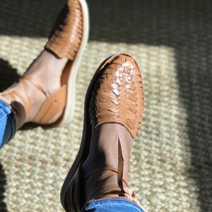 Puede incluir: Un par de sandalias de cuero marr&oacute;n con correas y cordones tejidos. Las sandalias se llevan en los pies de una persona, con los cordones atados alrededor de los tobillos.