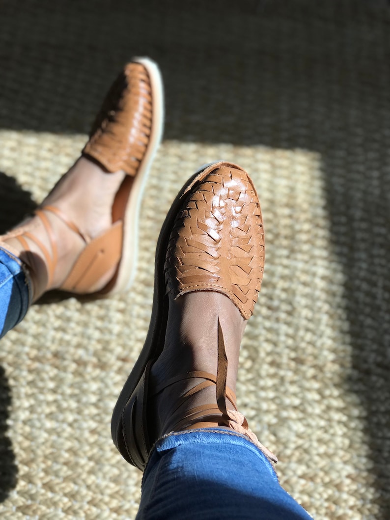 Puede incluir: Un par de sandalias de cuero marr&oacute;n con correas y cordones tejidos. Las sandalias se llevan en los pies de una persona, con los cordones atados alrededor de los tobillos.