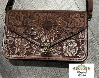 May include: A brown leather crossbody bag with a floral design. The bag features a sunflower and rose pattern, a metal clasp, and a long strap. The bag is from Magical Mexico, an artisanal boutique.