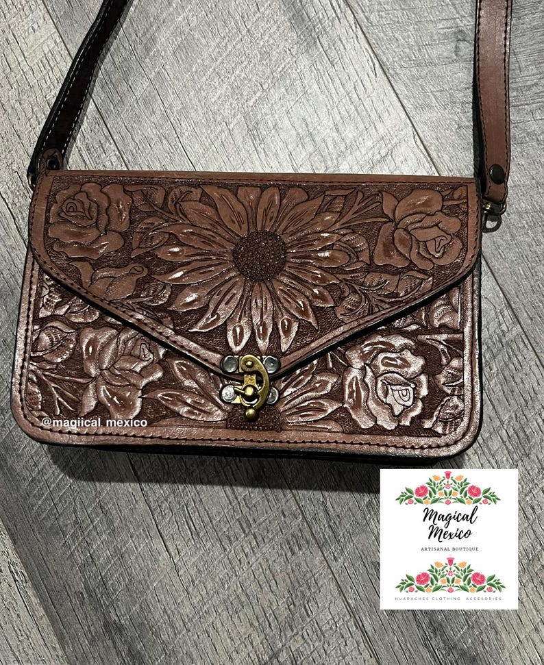 May include: A brown leather crossbody bag with a floral design. The bag features a sunflower and rose pattern, a metal clasp, and a long strap. The bag is from Magical Mexico, an artisanal boutique.