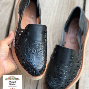 Peut inclure: Paire de huaraches en cuir noir avec un motif floral en relief sur l'empeigne. Les chaussures ont des c&ocirc;t&eacute;s tress&eacute;s, une semelle beige et des coutures blanches. Image prise sur une surface en bois.