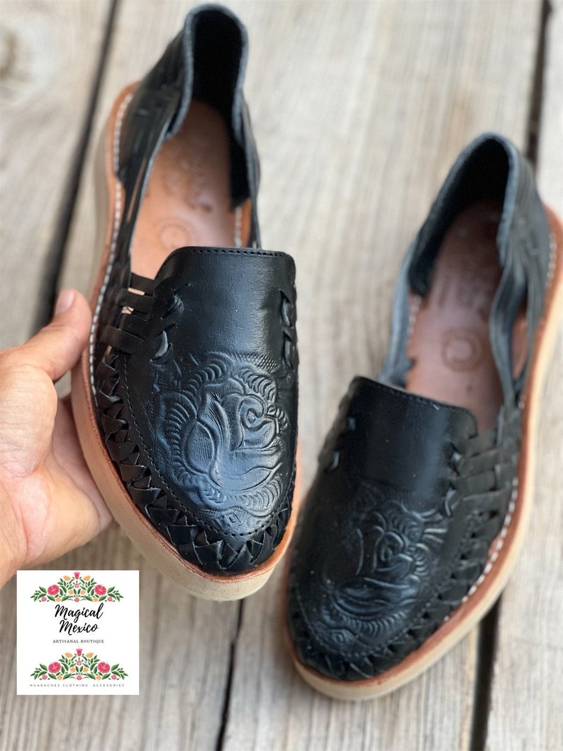 Peut inclure: Paire de huaraches en cuir noir avec un motif floral en relief sur l'empeigne. Les chaussures ont des c&ocirc;t&eacute;s tress&eacute;s, une semelle beige et des coutures blanches. Image prise sur une surface en bois.