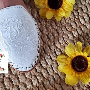 K&ouml;nnte beinhalten: Wei&szlig;e Ledersandalen mit Blumenmuster. Die Sandalen befinden sich auf einem gewebten Hintergrund mit gelben Sonnenblumen.
