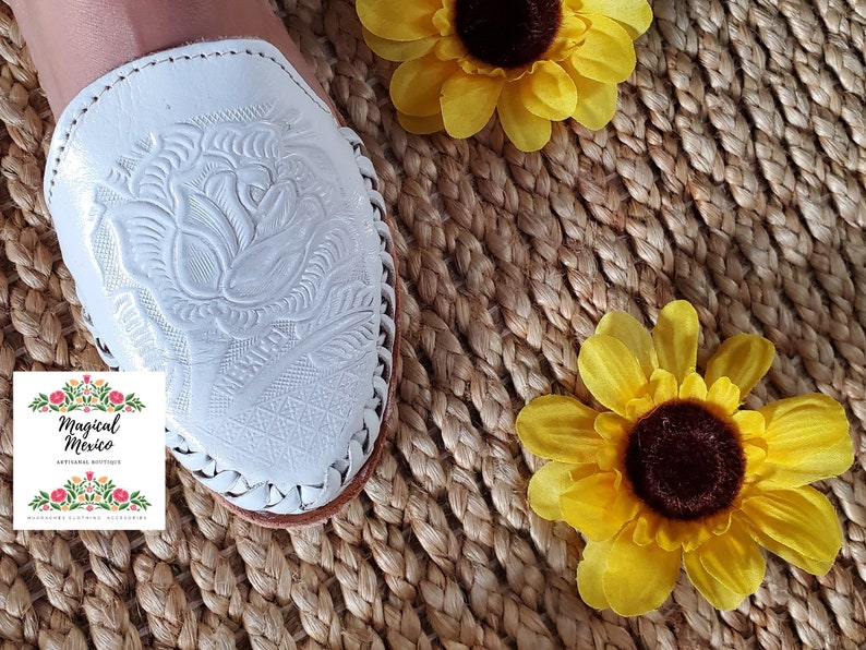 K&ouml;nnte beinhalten: Wei&szlig;e Ledersandalen mit Blumenmuster. Die Sandalen befinden sich auf einem gewebten Hintergrund mit gelben Sonnenblumen.