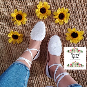 K&ouml;nnte beinhalten: Wei&szlig;e Ledersandalen mit Kn&ouml;chelriemen und aufwendigen Blumenstickereien. Die Sandalen befinden sich auf einem gewebten Hintergrund mit gelben Sonnenblumen.