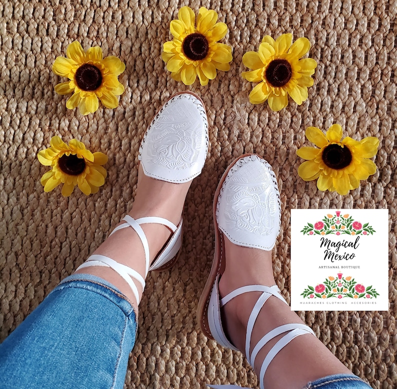 K&ouml;nnte beinhalten: Wei&szlig;e Ledersandalen mit Kn&ouml;chelriemen und aufwendigen Blumenstickereien. Die Sandalen befinden sich auf einem gewebten Hintergrund mit gelben Sonnenblumen.